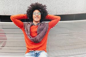 une Beau Jeune égyptien frisé gars séance sur une banc habillé dans une brillant Orange chandail et jeans. content personnes. le étudiant des promenades dans le Pause entre le cours. photo