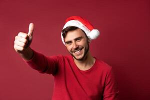 Jeune Beau caucasien gars dans une rouge chandail et Père Noël Chapeaux des stands sur rouge Contexte dans studio et montrant les pouces en haut. photo