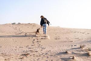 une Jeune caucasien fille quelque part dans une cuir veste et bleu jeans court le long de le sablonneux plage avec sa beagle chien photo