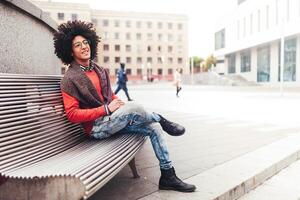 une Beau Jeune égyptien frisé gars séance sur une banc habillé dans une brillant Orange chandail et jeans. content personnes. le étudiant des promenades dans le Pause entre le cours. photo