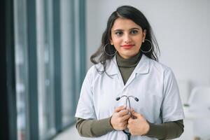 souriant femelle médecin posant dans hôpital bureau. content Jeune Indien femme médecin portant blanc médical manteau et stéthoscope à la recherche à caméra photo