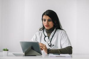 Indien femelle médecin dans une blanc manteau avec une stéthoscope conduit un en ligne consultation dans sa bureau. femelle médecin communique avec le téléspectateur directement dans le caméra photo