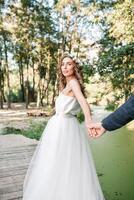 suivre moi mon l'amour concept. attrayant Jeune femme habillé dans blanc mariage robe en portant main de sa copain et en marchant sur le magnifique jardin à content avenir. photo