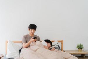 couple gay asiatique utilisant un téléphone portable à la maison. jeune homme asiatique lgbtq heureux se détendre après le réveil, vérifier les médias sociaux pendant que son petit ami dort allongé sur le lit dans la chambre à la maison le matin concept. photo