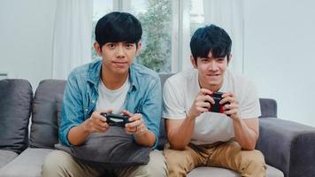 un jeune couple gay asiatique joue à des jeux à la maison, des adolescents coréens lgbtq utilisant un joystick ayant un moment de bonheur amusant ensemble sur un canapé dans le salon de la maison.