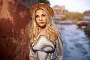 une magnifique Jeune blond femelle étudiant dans une paille chapeau et une rayé chemisier est permanent seul sur le rue. le concept de solitude et tristesse photo