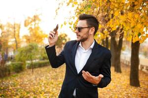 Beau Jeune élégant Masculin homme d'affaire très surpris par désagréable téléphone appel sur une marcher dans le l'automne parc sur le Contexte de Jaune feuilles photo