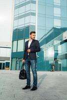 Beau Jeune homme d'affaire avec une barbe et dans une affaires costume permanent sur le rue contre le Contexte de le Bureau bâtiment suivant à une confortable élégant cuir sac. photo