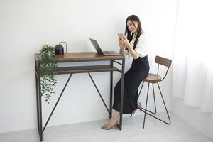 une Japonais femme vérification téléphone intelligent par éloigné travail dans le Accueil Bureau photo