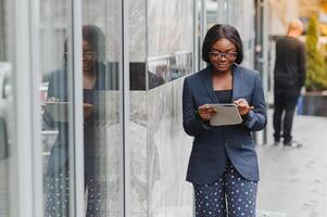 sur de soi femme d'affaires dehors. noir femme dans Bureau costume permanent près Urbain verre mur et en utilisant mobile téléphone. africain américain affaires femme concept photo