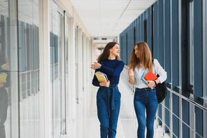 deux Jeune femmes avec livre bavardage tandis que permanent dans Université couloir. Université élèves dans couloir après le conférence. photo