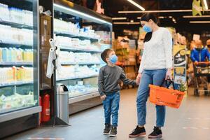 achats avec enfant pendant virus épidémie. mère et enfant portant chirurgical visage masque achat fruit dans supermarché. maman et peu garçon acheter Frais légume dans épicerie magasin. famille dans magasin photo