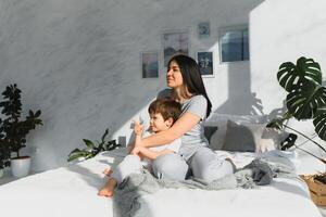 Jeune mère avec sa 5 ans vieux peu fils habillé dans pyjamas sont relaxant et en jouant dans le lit à le fin de semaine ensemble, paresseux matin, chaud et confortable scène. famille Accueil tirer. photo