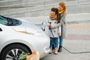 une élégant mère enseigne sa fille Comment à correctement charge un électrique auto. photo