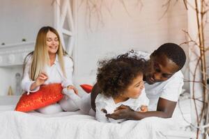 portrait de content multiracial Jeune famille mensonge sur confortable blanc lit à maison, souriant international maman et papa relaxant avec peu biracial fille enfant posant pour image dans chambre photo