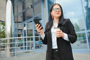 avocat femme d'affaires professionnel en marchant en plein air parlant sur cellule intelligent téléphone en buvant café de jetable papier Coupe. photo