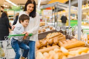 mignonne enfant avec mère choisir cuit des biens ou Pâtisserie dans boulangerie magasin. peu garçon avec maman achat pain ou petits pains à épicerie supermarché photo