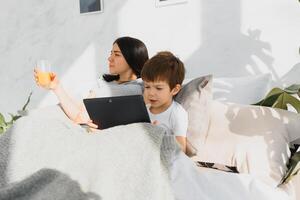 magnifique souriant mère et fils en jouant avec numérique tablette pose sur lit. photo