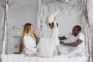 portrait de content multiracial Jeune famille mensonge sur confortable blanc lit à maison, souriant international maman et papa relaxant avec peu biracial fille enfant posant pour image dans chambre photo