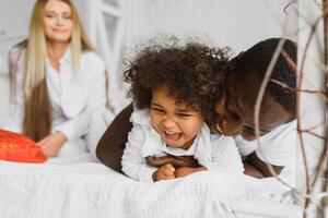 portrait de content multiracial Jeune famille mensonge sur confortable blanc lit à maison, souriant international maman et papa relaxant avec peu biracial fille enfant posant pour image dans chambre photo