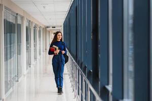 de bonne humeur brunette étudiant fille avec noir sac à dos détient livres dans moderne bâtiment. femelle étudiant permanent avec livres dans Université couloir photo