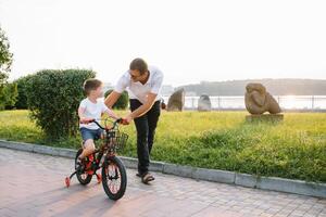 Jeune et content père enseigne le sien Jeune fils à balade une vélo. le enfant est content . père en train de regarder fils. du père journée photo