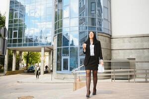 Jeune fille Bureau ouvrier dans lunettes, foncé veste et bleu chemise séjours sur ville rue près Bureau bâtiment et détient sa documents. photo