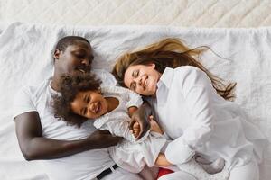 portrait de content multiracial Jeune famille mensonge sur confortable blanc lit à maison, souriant international maman et papa relaxant avec peu biracial fille enfant posant pour image dans chambre photo