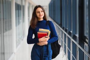 de bonne humeur brunette étudiant fille avec noir sac à dos détient livres dans moderne bâtiment. femelle étudiant permanent avec livres dans Université couloir photo