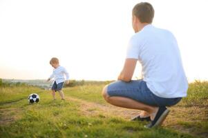 père avec une peu fils pièces Football sur le vert herbe dans le parc. content famille ayant amusement et en jouant Football sur une vert herbeux pelouse sur une ensoleillé journée. famille concept, du père journée photo