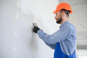 homme cloison sèche ouvrier ou plâtrier ponçage et lissage une placoplâtre des murs avec stuc en utilisant une papier de verre titulaire. portant blanc casque et sécurité lunettes. panoramique image avec copie espace photo