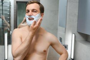 Jeune souriant caucasien torse nu homme appliquant rasage mousse sur visage dans de face de miroir, en train de préparer pour cheveux suppression Matin routine. concept de personnel hygiène, soins de la peau, Masculin faciale beauté des produits. photo