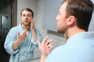 Beau homme à la recherche à miroir et appliquant hydratant crème sur des joues dans salle de bains. soigné Jeune gars Faire soin de la peau Matin routine après prise une douche. photo