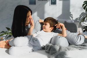 Jeune mère avec sa 4 ans vieux peu fils habillé dans pyjamas sont relaxant et en jouant dans le lit à le fin de semaine ensemble, paresseux matin, chaud et confortable scène. photo