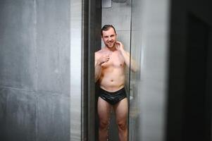 Beau Jeune homme prise une douche permanent en dessous de chute l'eau gouttes la lessive nu corps et tête dans moderne salle de bains à maison. Masculin modèle photo