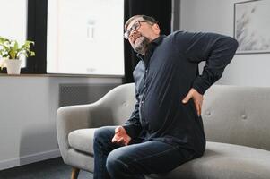 personnes, soins de santé et traitement. portrait de malade mature homme Souffrance de aigu côté retour douleur, séance sur canapé à maison, la personne émouvant inférieur retour douloureux zone avec main. santé problème concept photo