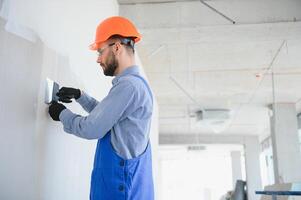 homme cloison sèche ouvrier ou plâtrier ponçage et lissage une placoplâtre des murs avec stuc en utilisant une papier de verre titulaire. portant blanc casque et sécurité lunettes. panoramique image avec copie espace photo