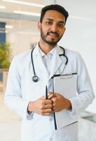 portrait de content amical Masculin Indien Latin médecin médical ouvrier portant blanc manteau avec stéthoscope autour cou permanent dans moderne privé clinique à la recherche à caméra. médical soins de santé concept photo