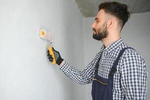 Jeune souriant professionnel ouvrier dans bleu uniforme permanent avec peindre rouleau dans Nouveau appartement pour réparer plus de gris des murs arrière-plan, copie espace. photo