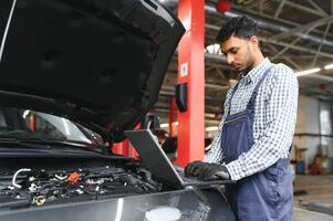 mécanicien homme mécanicien directeur ouvrier en utilisant une portable ordinateur vérification voiture dans atelier à auto voiture réparation un service centre. ingénieur Jeune homme à la recherche à inspection véhicule détails en dessous de voiture capot. photo