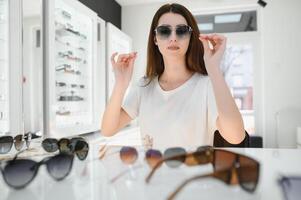 elle fabriqué sa choix. magnifique Jeune femme ajustement sa Nouveau des lunettes de soleil et souriant tandis que permanent dans optique magasin. photo