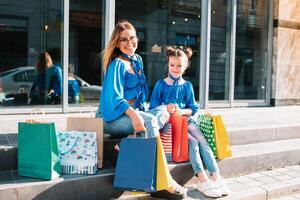 magnifique maman et sa mignonne peu fille sont en portant achats Sacs, à la recherche à caméra et souriant tandis que permanent en plein air. achats concept. photo