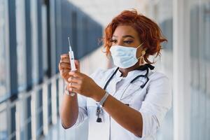 Jeune souriant africain américain médecin dans médical masque en portant une seringue photo