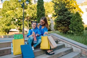 magnifique maman et sa mignonne peu fille sont en portant achats Sacs, à la recherche à caméra et souriant tandis que permanent en plein air. achats concept photo