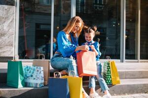 magnifique maman et sa mignonne peu fille sont en portant achats Sacs, à la recherche à caméra et souriant tandis que permanent en plein air. achats concept. photo