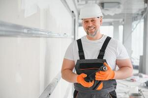 homme cloison sèche ouvrier ou plâtrier en mettant engrener ruban pour placoplâtre sur une mur en utilisant une spatule et plâtre. portant blanc casque, travail gants et sécurité lunettes. image avec copie espace. photo