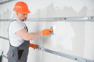 homme cloison sèche ouvrier ou plâtrier en mettant engrener ruban pour placoplâtre sur une mur en utilisant une spatule et plâtre. portant blanc casque, travail gants et sécurité lunettes. image avec copie espace. photo