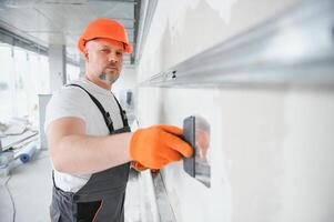 homme cloison sèche ouvrier ou plâtrier en mettant engrener ruban pour placoplâtre sur une mur en utilisant une spatule et plâtre. portant blanc casque, travail gants et sécurité lunettes. image avec copie espace. photo