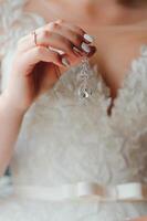 magnifique la mariée dans blanc mariage robe met sur boucle d'oreille. beauté modèle fille est portant bijoux. femelle portrait dans de mariée robe pour mariage. femme avec frisé cheveux et dentelle voile. mignonne Dame à l'intérieur photo