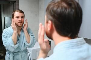 Beau homme à la recherche à miroir et appliquant hydratant crème sur des joues dans salle de bains. soigné Jeune gars Faire soin de la peau Matin routine après prise une douche. photo